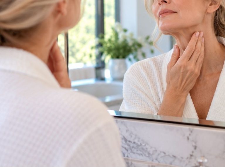 woman examining signs of neck aging and loose skin in a bathroom mirror, highlighting neck rejuvenation and deep plane neck lift procedures.