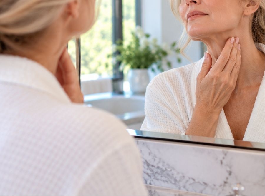 woman examining signs of neck aging and loose skin in a bathroom mirror, highlighting neck rejuvenation and deep plane neck lift procedures.